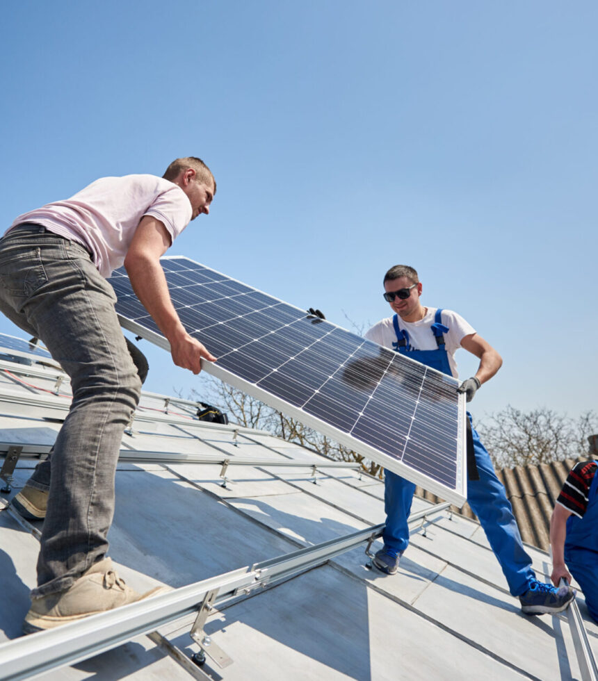 Male engineers installing solar photovoltaic panel system. Three electricians lifting blue solar module on roof of modern house. Alternative energy sustainable resources ecology concept.